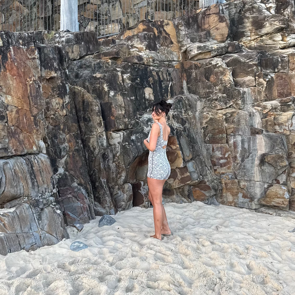 A woman stands on a sandy beach in front of a large rock wall, wearing a light-colored patterned romper or dress. A wooden walkway and metal railing are visible above the rocks.