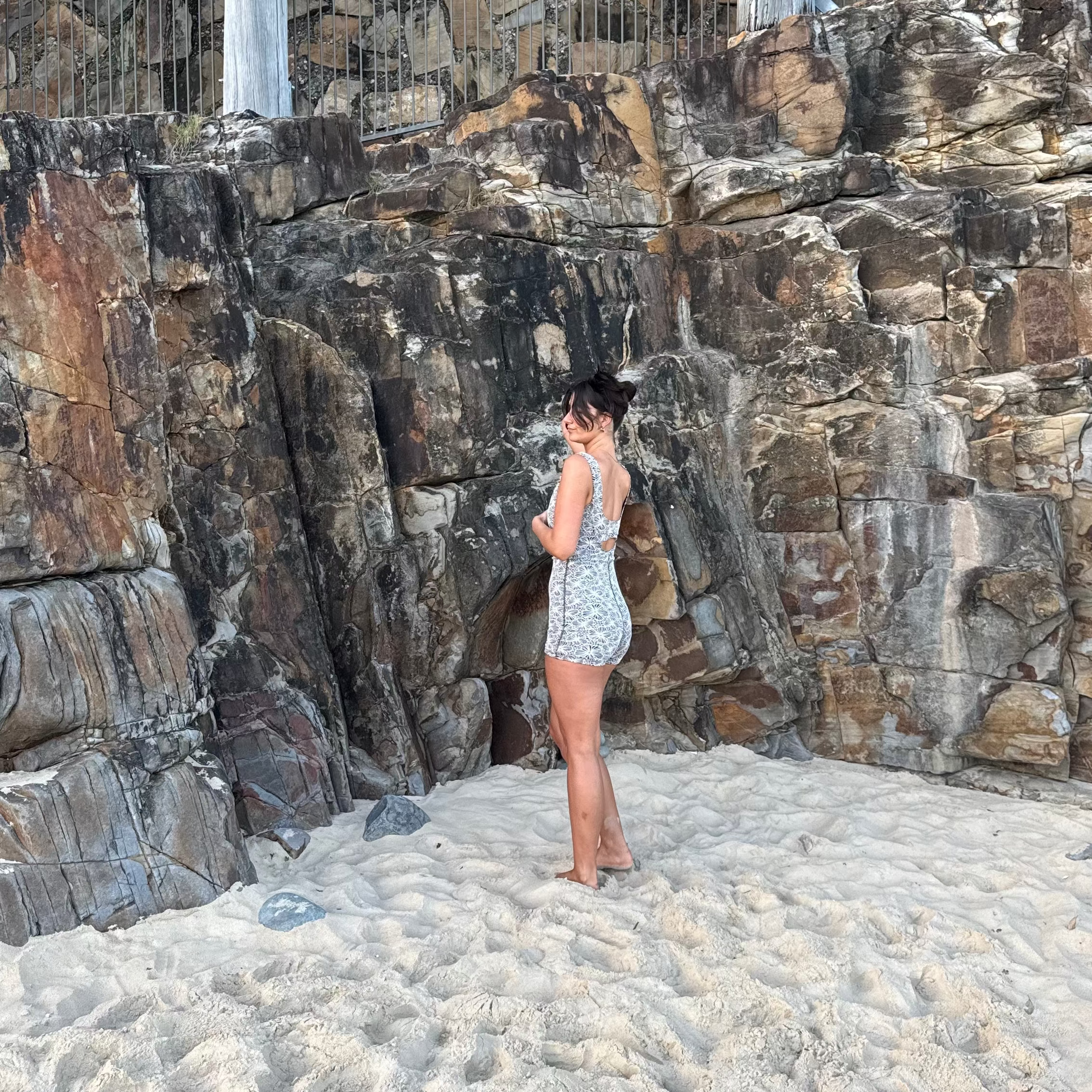 A woman stands on a sandy beach in front of a large rock wall, wearing a light-colored patterned romper or dress. A wooden walkway and metal railing are visible above the rocks.