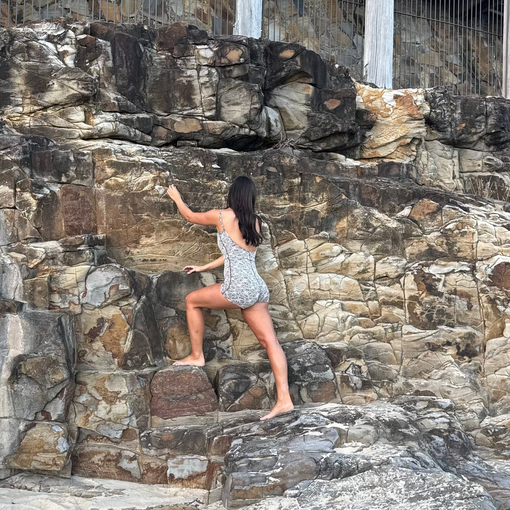 A woman in a patterned romper climbing on a large rock face outdoors, with sand in the foreground and a wooden structure above.