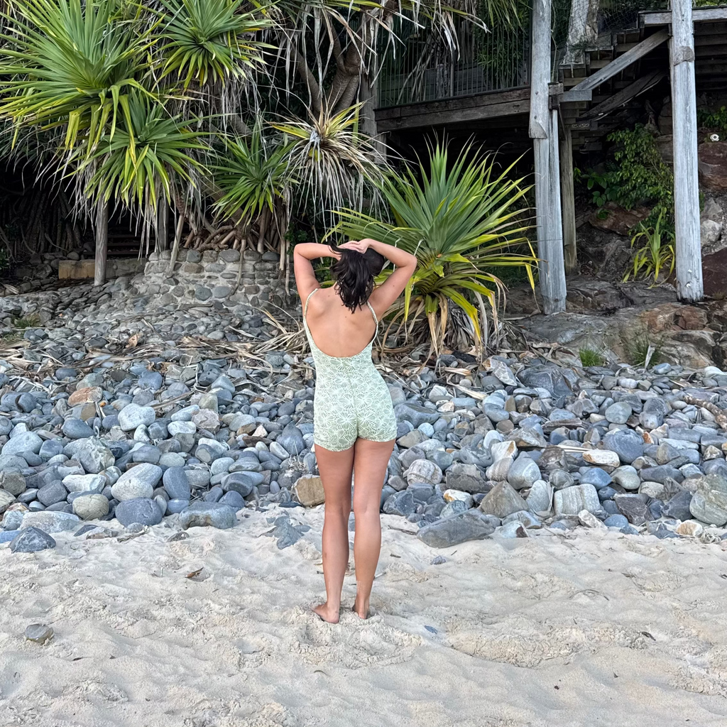A woman stands on a sandy beach with her back to the camera, wearing a light green and white patterned one-piece swimsuit. Behind her are rocky shores, lush green tropical plants, and a wooden walkway structure.